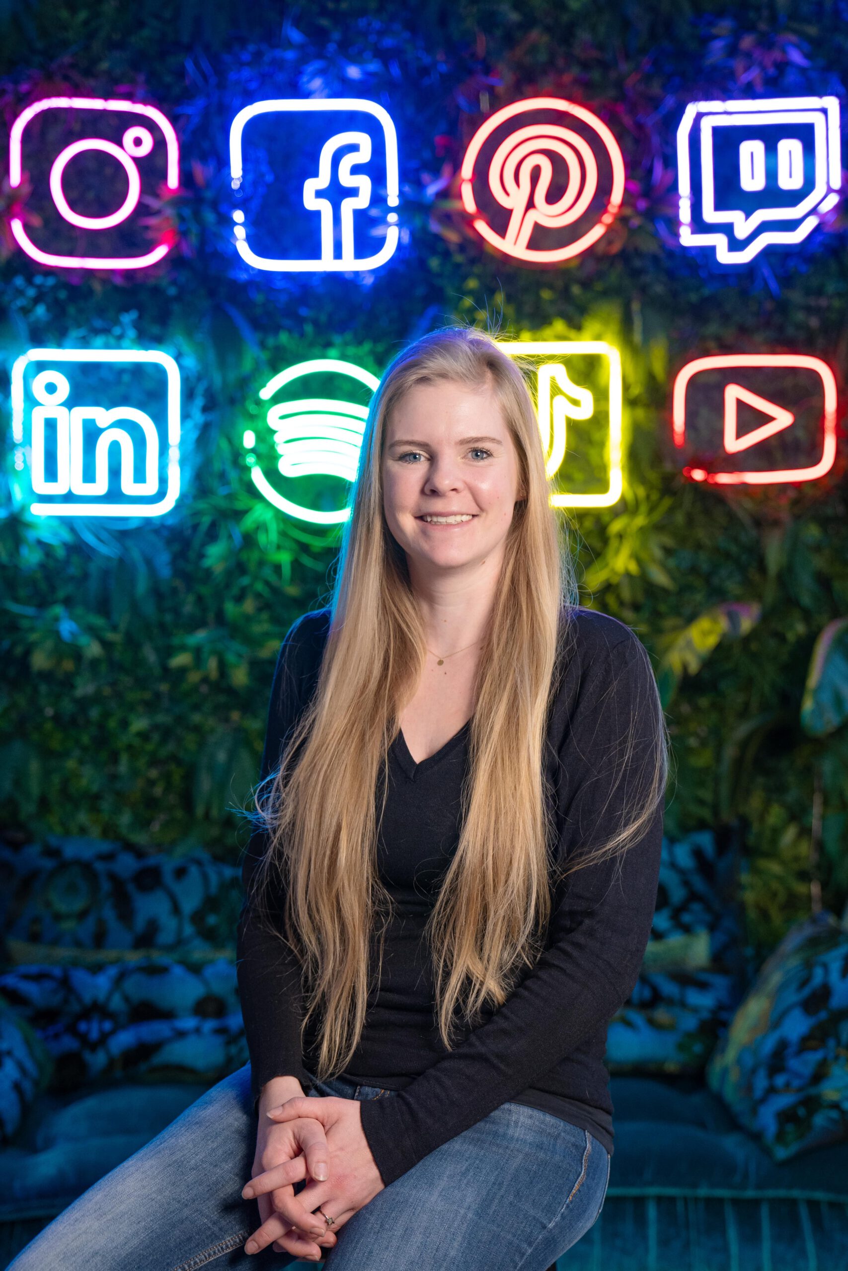 Junge blonde Frau mit sehr langen Haaren vor begrünter Wand mit Neon-Lichtern in Form von Social-Media-Icons